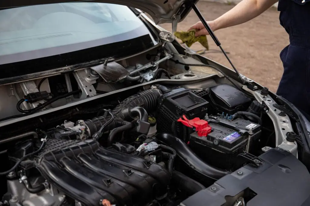 Open car hood showing the engine bay and battery, with a person beside the vehicle