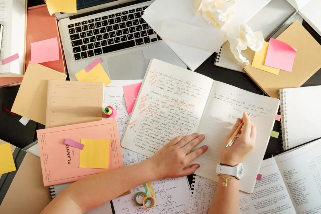 Hands writing notes at a desk with a laptop and study materials